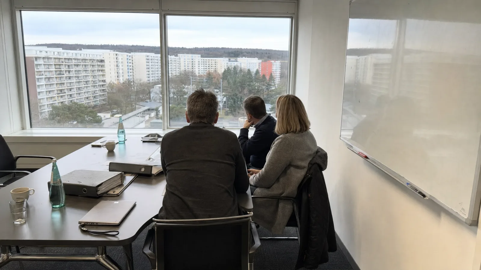 Besprechungsraum mit Aktenordnern, drei Personen blicken aus dem Fenster auf eine Wohnanlage