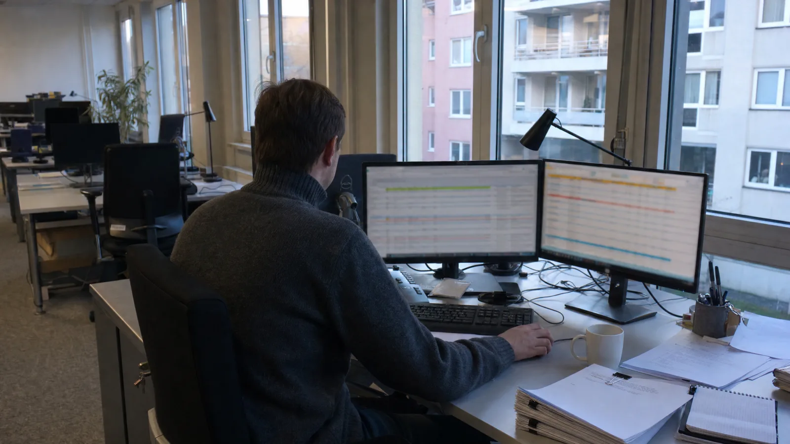 Schreibtisch am Fenster am frühen Morgen, eine Person arbeitet an zwei Bildschirmen